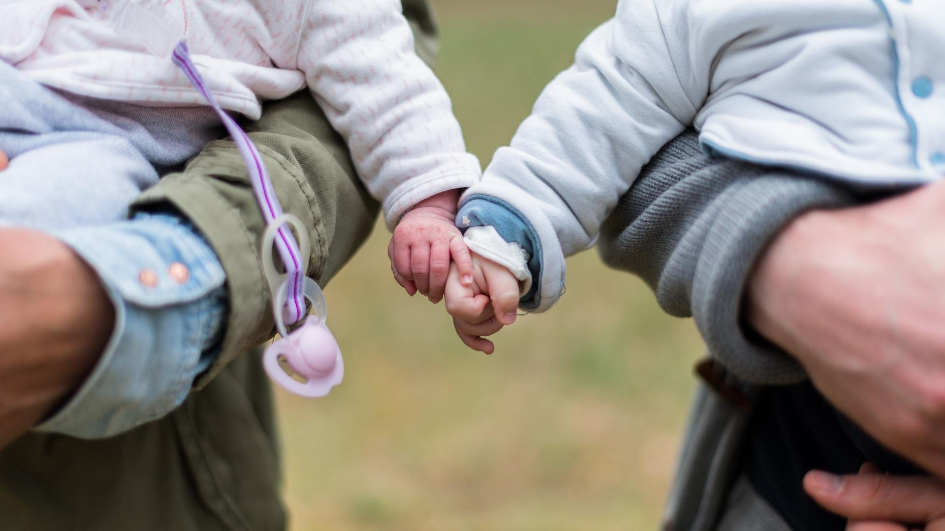 Stimmungsbild zum Beitrag: Es werden wwei Elternteile mit Babies im Arm abgebildet. Die Babies halten Händchen.