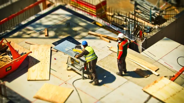 Stimmungsbild zum Beitrag: Zwei Männer auf einer Großbaustelle, die an einer Säge arbeiten, werden abgebildet.