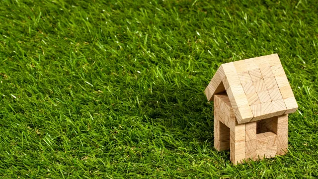Stimmungsbild zum Beitrag: Ein kleines Modelhaus, das aus hölzernen Bauklötzen gebaut ist, wird abgebildet. Es steht auf einer Grasfläche.