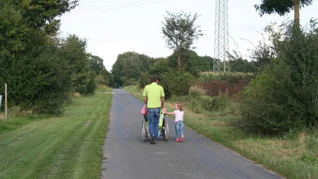 Stimmungsbild zum Beitrag: Es wird ein Erwachsener mit seinem daneben gehenden Kind und einer Person im Rollstuhl abgebildet.