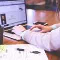 Stimmungsbild zum Beitrag: Eine Frau tippt am Laptop und neben ihr liegen Dokumente, ein Stift und ein Teebecher.