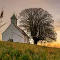 Stimmungsbild zum Beitrag: Es wird eine weiße Kapelle an einem kleinen Abhang mit einem daneben stehenden Baum abgebildet.