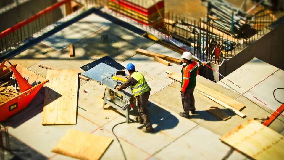 Stimmungsbild zum Beitrag: Zwei Männer auf einer Großbaustelle, die an einer Säge arbeiten, werden abgebildet.