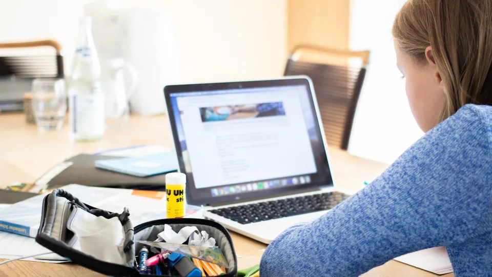 Stimmungsbild zum Beitrag: Ein Kind sitzt am Laptop und liest. Auf dem Tisch liegt neben ihr ein offenes Schulmäppchen.