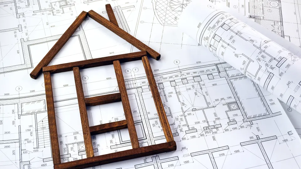 Stimmungsbild zum Beitrag: Es ist ein mit Holzblöcken zusammengesetztes Haus auf einem Bauplan abgebildet.