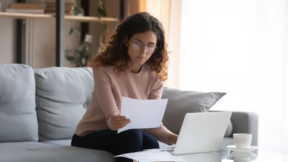 Stimmungsbild zum Beitrag: Eine Frau sitzt auf einem Sofa und hat ein Blatt Papier in der Hand. Auf dem Tisch vor ihr liegt ein Laptop.